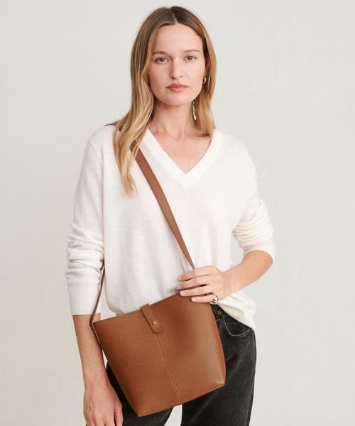A woman with long, light brown hair wears a white V-neck sweater and black pants while holding the Mini Leather Bucket Bag, handmade in Italy. She stands against a plain light background, gazing calmly at the camera.