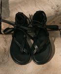 The Nina Sandal, featuring black crisscross straps and leather laces, rests on a textured stone surface in the sunlight.