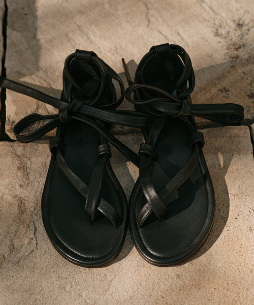 The Nina Sandal, featuring black crisscross straps and leather laces, rests on a textured stone surface in the sunlight.