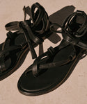 A pair of Nina Sandal black leather sandals featuring crisscross straps and ankle ties, displayed on a light beige surface in natural sunlight.