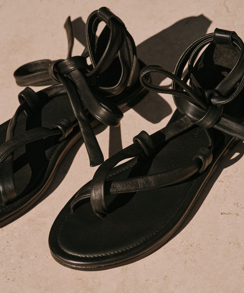 A pair of Nina Sandal black leather sandals featuring crisscross straps and ankle ties, displayed on a light beige surface in natural sunlight.
