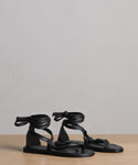 A pair of Nina Sandal black sandals with leather laces that wrap around the ankle and tie in a bow, shown on a wooden surface with a plain light gray background.