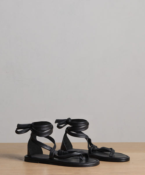A pair of Nina Sandal black sandals with leather laces that wrap around the ankle and tie in a bow, shown on a wooden surface with a plain light gray background.