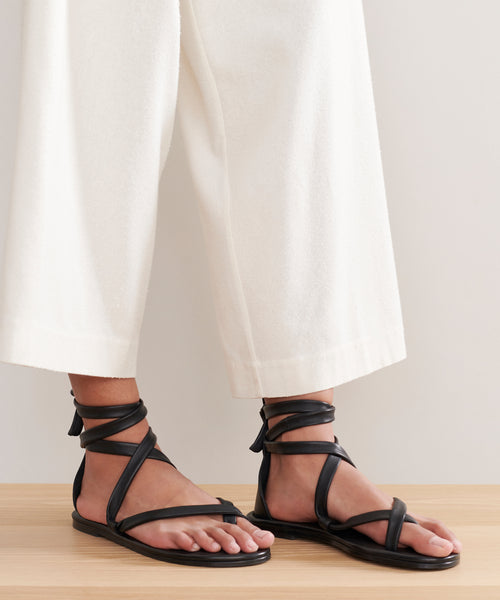 Person wearing off-white, wide-leg, cropped pants and the Nina Sandal featuring black leather laces and ankle ties, standing on a light wooden surface.