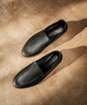 A pair of Palma Loafer shoes in black Italian leather rests on a light brown textured surface, photographed from above with soft natural light creating elegant shadows.