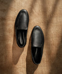 A pair of Palma Loafer black leather slip-ons is displayed on a textured brown surface, illuminated by gentle natural light and shadows.