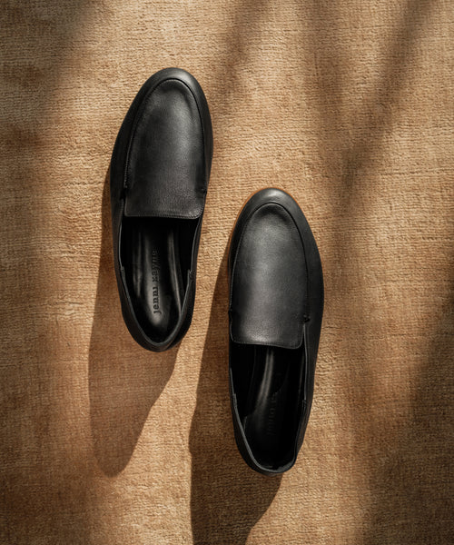 A pair of Palma Loafer black leather slip-ons is displayed on a textured brown surface, illuminated by gentle natural light and shadows.