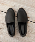 A pair of Palma Loafers in black Italian leather, featuring visible stitching, displayed side by side on a rustic wooden surface.