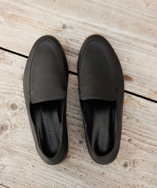 A pair of Palma Loafers in black Italian leather, featuring visible stitching, displayed side by side on a rustic wooden surface.