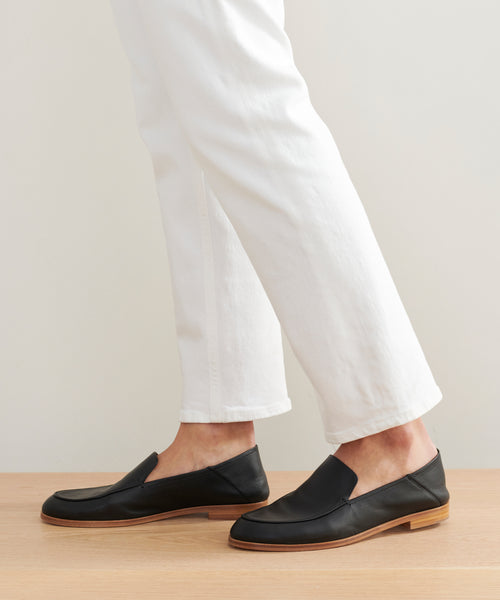 A person in white pants wears black Palma Loafer Italian leather shoes with light brown soles, standing on a wooden surface against a neutral background.