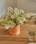 A bouquet of white flowers in the Small Leather Rivet Vase rests on a wooden table arranged with clear glasses of water and plates, all bathed in soft natural light.