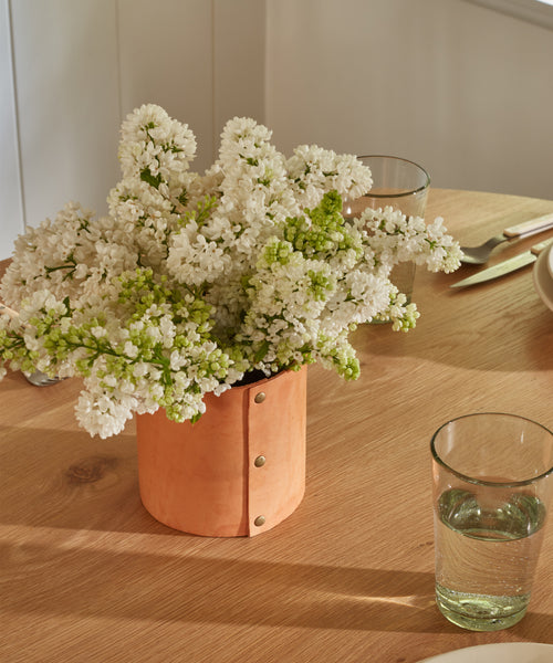 A bouquet of white flowers in the Small Leather Rivet Vase rests on a wooden table arranged with clear glasses of water and plates, all bathed in soft natural light.