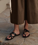 A person stands on stone wearing the Scarlett Sandal—black Italian leather sandals with crisscross straps—paired with a long brown skirt for an effortlessly chic summer look.