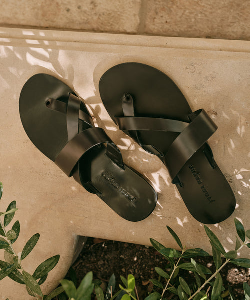 The Scarlett Sandal, a pair of black leather sandals with crisscross straps, made in Italy, rests on a light stone surface amid green plants and dappled sunlight.