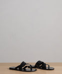 The Scarlett Sandal, a pair of black leather summer sandals with crisscross straps, is displayed on a light wooden surface against a plain, light background.