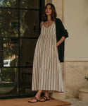 A woman stands by a glass door in a long, sleeveless cream dress with black stripes, a dark cardigan over her shoulders, and stylish Scarlett Sandals made in Italy. She looks ahead with one hand in her pocket.