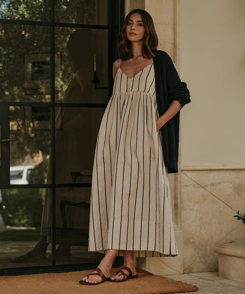 A woman stands by a glass door in a long, sleeveless cream dress with black stripes, a dark cardigan over her shoulders, and stylish Scarlett Sandals made in Italy. She looks ahead with one hand in her pocket.