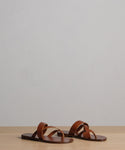 The Scarlett Sandal, a pair of brown leather sandals with crisscross straps made in Italy, is displayed on a light wooden surface against a plain light gray background.