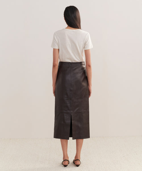 A woman with long straight hair faces away, wearing a white short-sleeve tee tucked into the high-waisted Leather Sienna Skirt in chocolate with a back slit. She has black shoes. The background is plain.