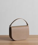 The Leather Sloane Bag in taupe pebbled Epsom leather, with a curved flap and single top handle, is displayed on a light wooden surface against a plain light gray backdrop.