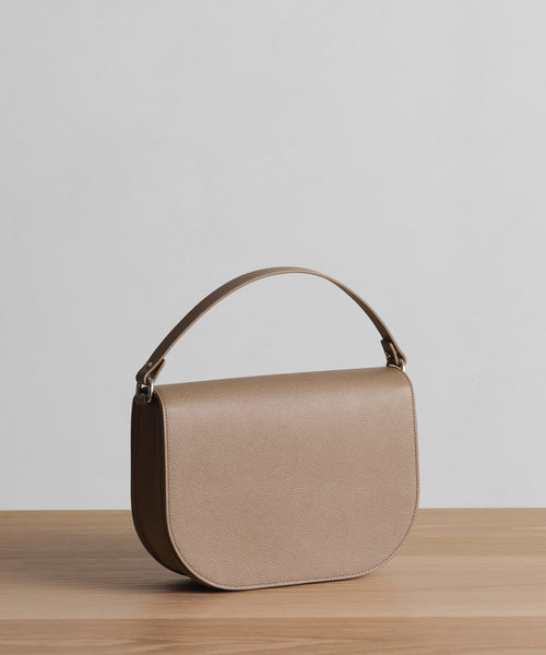The Leather Sloane Bag in taupe pebbled Epsom leather, with a curved flap and single top handle, is displayed on a light wooden surface against a plain light gray backdrop.