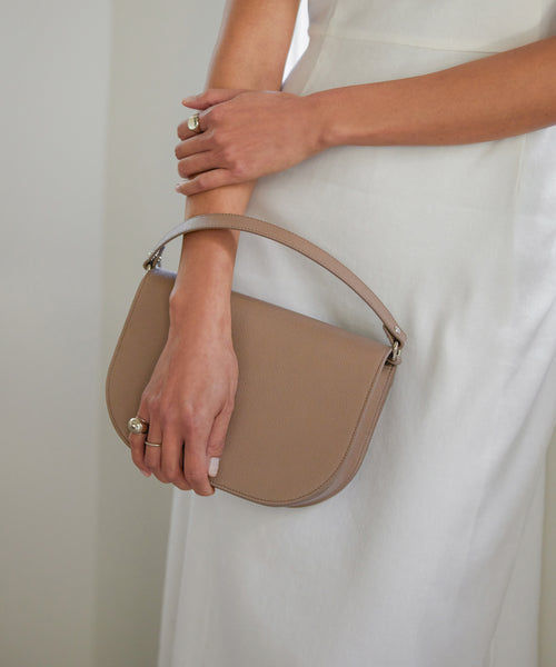 A person in a white dress, arms crossed and rings on their fingers, holds the taupe Leather Sloane Bag with a half-moon silhouette by its handle. The bag features pebbled epsom leather for timeless elegance.