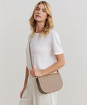A woman with wavy blonde hair, dressed in a white t-shirt and light pants, stands against a plain background looking at the camera. She carries the Leather Sloane Bag over her shoulder.