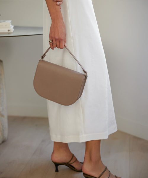 A person in a white dress and heeled sandals holds the Leather Sloane Bag by its top handle, standing indoors on a light wooden floor near a table.