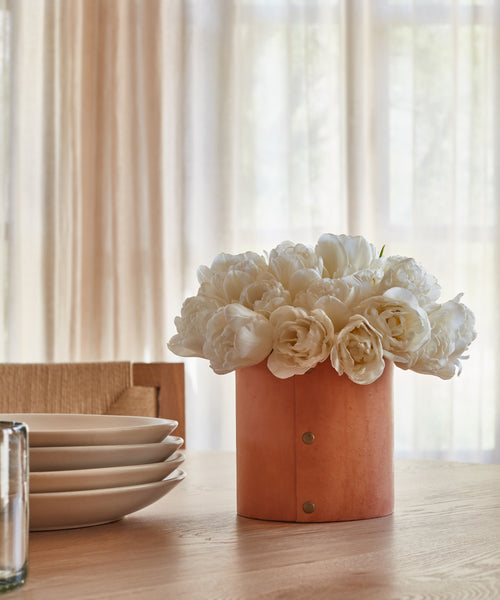 The Small Leather Rivet Vase, filled with white peonies, sits on a wooden table beside stacked white plates, with sheer curtains and gentle daylight in the background.