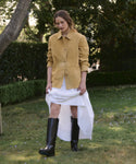 Outdoors on grass, a person wears a tan jacket, a long white dress, and the Leather Stowe Boot—tall black calf leather boots—standing amid trees and hedges.