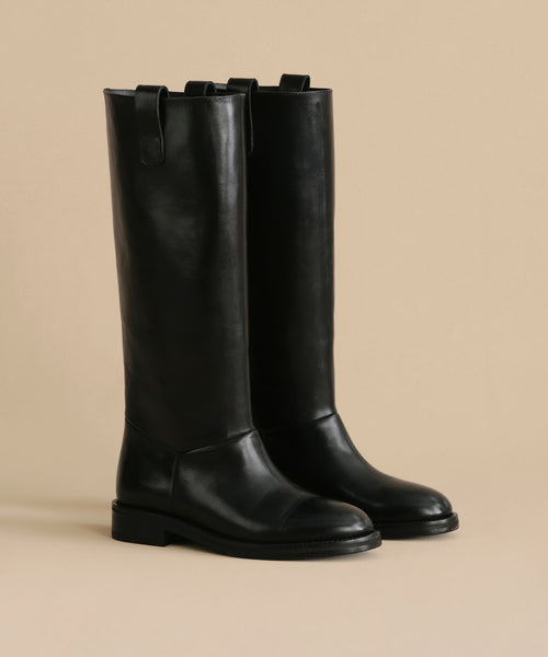 The Leather Stowe Boot is a pair of tall black calf leather riding boots with low heels and pull tabs, displayed upright against a light beige background.