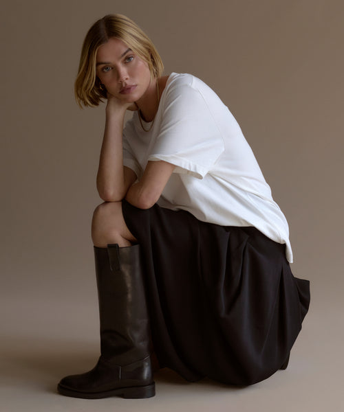 A woman with short blonde hair crouches, resting her chin on her hand. She wears a loose white t-shirt, black skirt, and tall Leather Stowe Boots crafted from calf leather, against a plain neutral background.