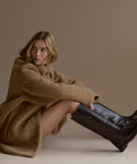 A woman with a blonde bob sits sideways on the floor, wearing a tan faux fur coat and black Leather Stowe Boots, against a plain beige background.