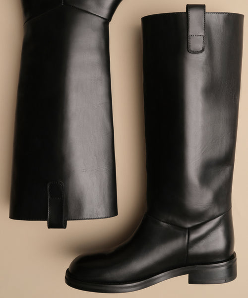 The Leather Stowe Boot, shown upright and on its side against a beige background, features smooth black calf leather, pull tabs at the top, and a minimalist design reminiscent of a classic riding boot.