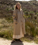 Outdoors on sandy ground, a person wears the Leo Trench Coat in dark khaki over a white pleated skirt and black sandals. Tall grasses and shrubs fill the sunlit background.
