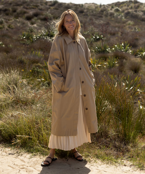 Outdoors on sandy ground, a person wears the Leo Trench Coat in dark khaki over a white pleated skirt and black sandals. Tall grasses and shrubs fill the sunlit background.