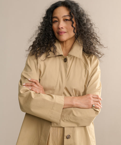 A woman with curly dark hair stands with her arms crossed, smiling softly against a neutral background while wearing the Leo Trench Coat.