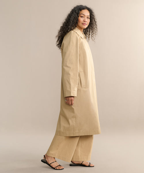 A person with long curly hair, dressed in the Leo Trench Coat, matching wide-leg pants, and black sandals, stands sideways against a light beige background.