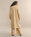 A person with long, curly hair stands facing away, wearing the dark khaki Leo Trench Coat with matching wide-leg pants and black heeled sandals, set against a neutral beige background.