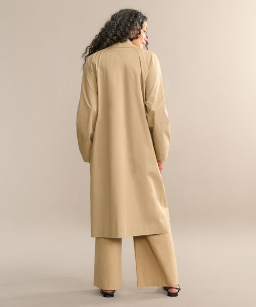 A person with long, curly hair stands facing away, wearing the dark khaki Leo Trench Coat with matching wide-leg pants and black heeled sandals, set against a neutral beige background.