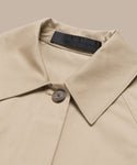 Close-up of the Leo Trench Coat—a minimalist design in dark khaki with a single brown button and black neckline label—displayed against a beige background.