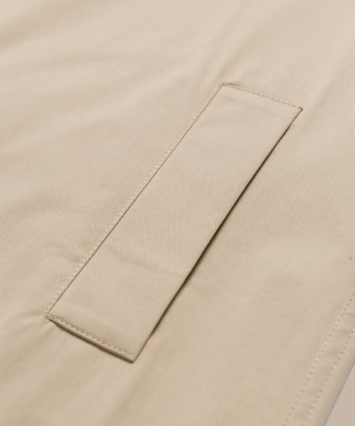 Close-up of a beige fabric with a neatly stitched rectangular flap, likely part of a pocket on the minimalist Leo Trench Coat.