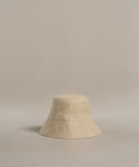 The Linen Bucket Hat, crafted in Portugal from beige linen, features a simple design and downward-sloping brim. Photographed on a neutral background, it’s the perfect minimalist accessory for summer beach days.