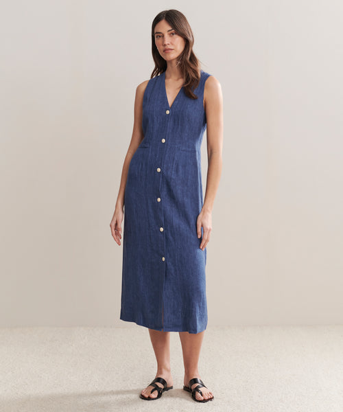 A woman with long brown hair and a neutral expression stands against a plain background wearing the Callan Dress, a sleeveless, button-down chambray style with a V-neckline, paired with black sandals.