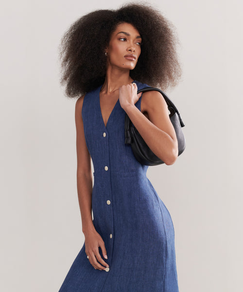 A woman with voluminous curly hair confidently poses against a plain background, wearing the sleeveless, button-down Callan Dress and carrying a black handbag over her shoulder.