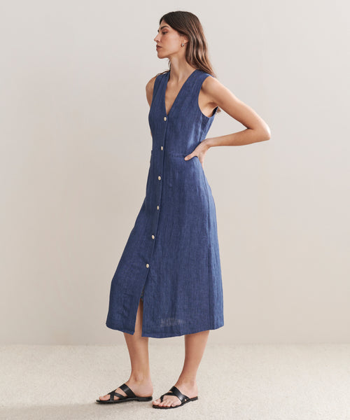 A woman stands sideways against a plain light background, wearing the Callan Dress—a sleeveless blue chambray midi vest dress with a V-neck—paired with black sandals.