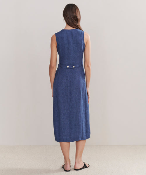 A woman with long brown hair stands facing away, wearing the sleeveless, knee-length Callan Dress with two buttons on the back waistband and black sandals, set against a plain light background.