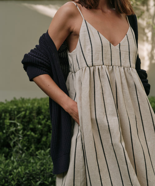 A woman stands outdoors with one hand in her pocket, wearing the relaxed fit Cove Dress in Natural/Navy Stripe and a dark cardigan over her shoulders. Lush greenery appears in the background.