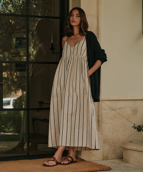 A woman stands indoors by a glass door, wearing the Cove Dress. The dress has a relaxed fit with vertical stripes and is styled with a black cardigan and brown sandals. She has one hand in her pocket and a neutral expression.