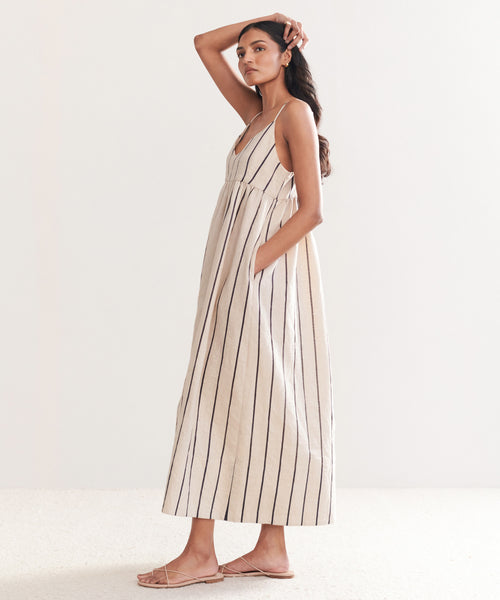 Woman with long dark hair models the Cove Dress, a sleeveless style with side pockets and a relaxed fit in Natural/Navy Stripe. She stands sideways in flat sandals against a plain light background.
